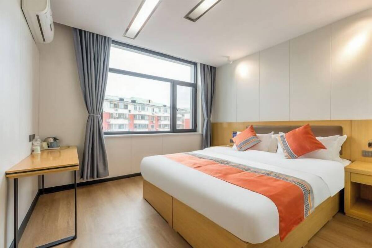 A hotel room with a bed, a desk, a window with curtains, and a view of buildings outside.
