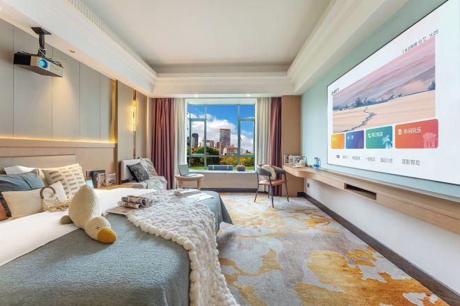 Habitación de hotel con una cama grande, un escritorio, una pantalla de proyector y vistas a la ciudad desde la ventana.