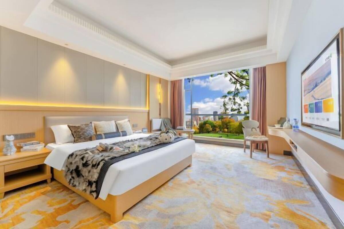 Una habitación de hotel con una cama grande, un escritorio, una silla, un ventanal con vistas a zonas verdes y un televisor montado en la pared.