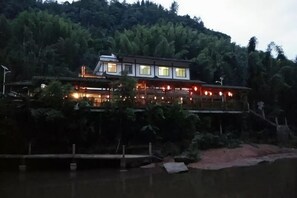 A multi-story building with a balcony, situated by a body of water, surrounded by dense greenery. - Puyuan Xiangu Hotel (Chishui)
