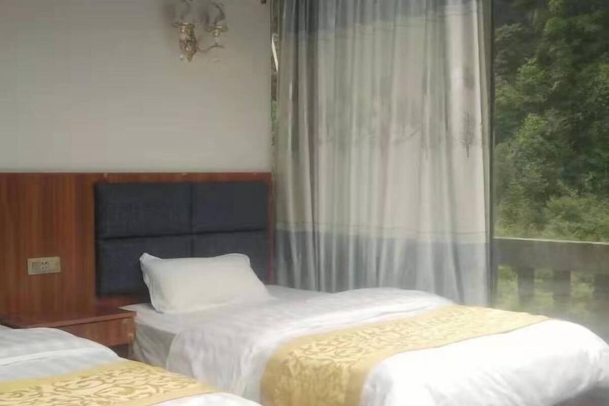 A hotel room with two beds, a wooden headboard, a window with sheer curtains, and a view of greenery outside.
