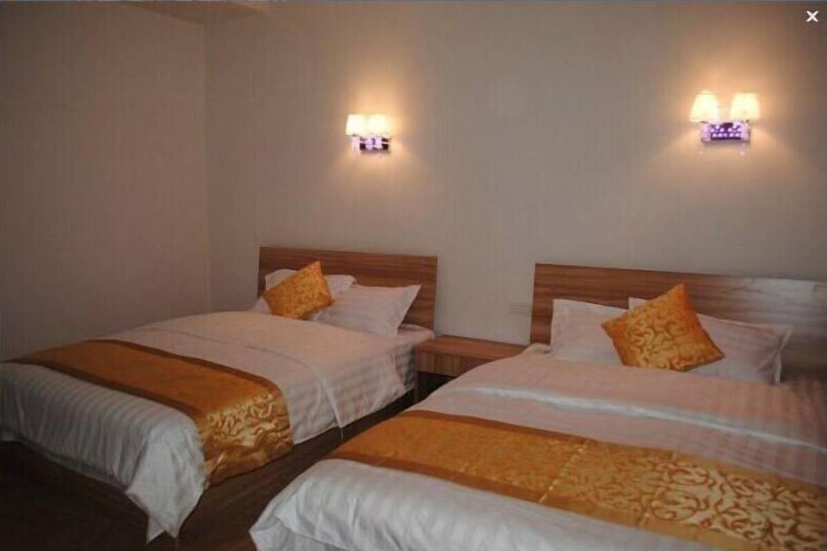 A hotel room with two beds, each with a wooden headboard, white bedding, and a patterned throw blanket. There are wall-mounted lamps on either side of the beds.