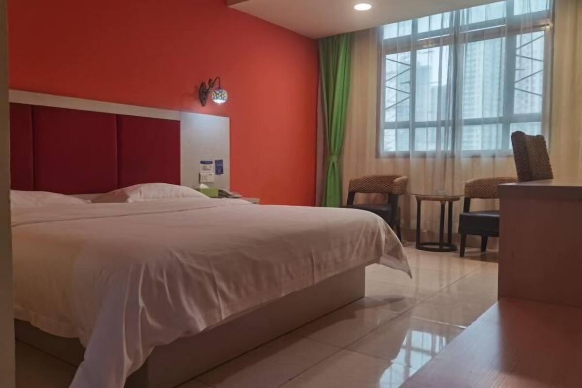 A hotel room with a large bed, a desk, a chair, a window with curtains, and a red wall.