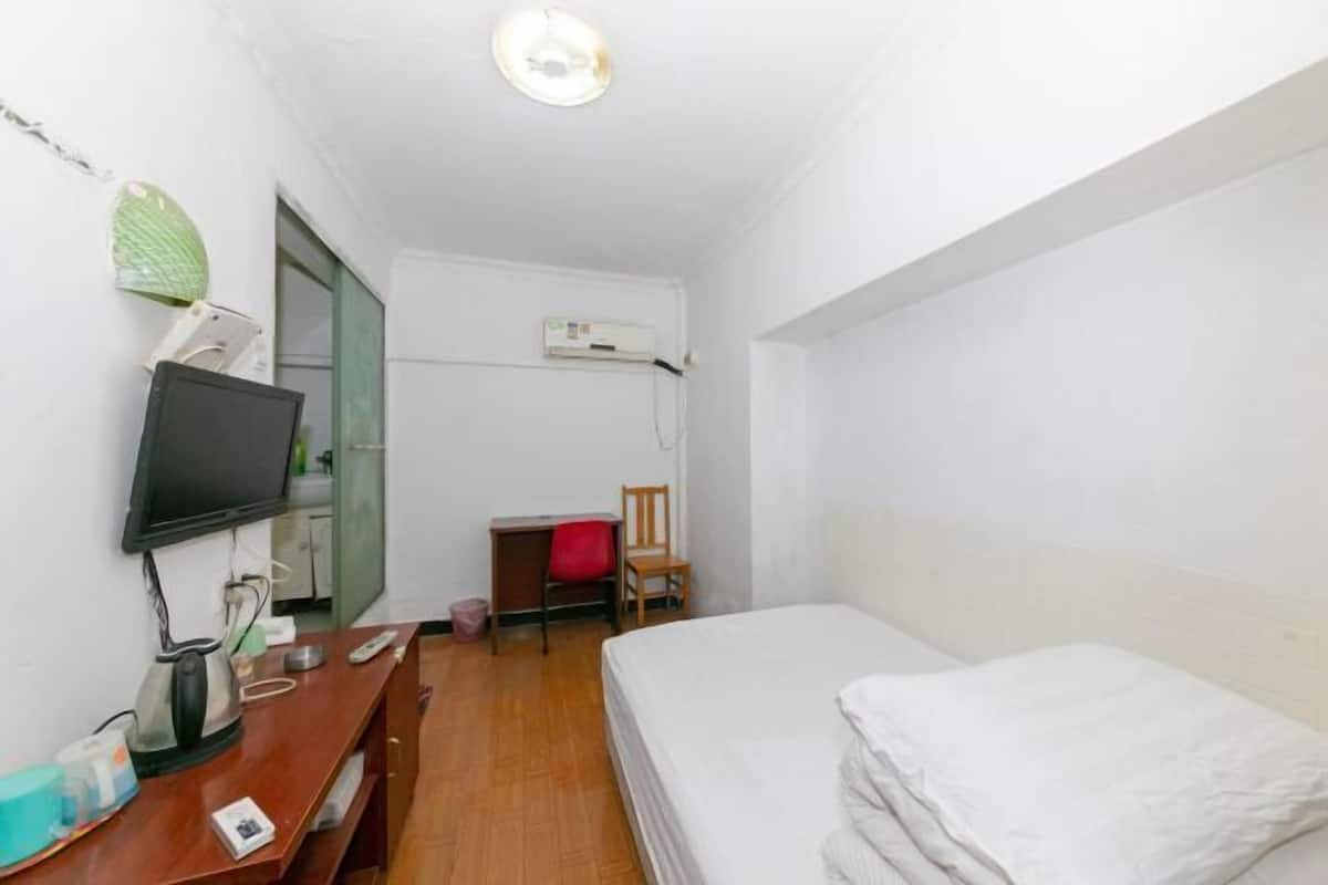A small, clean room with a bed, a desk, a chair, a TV, and an air conditioning unit.