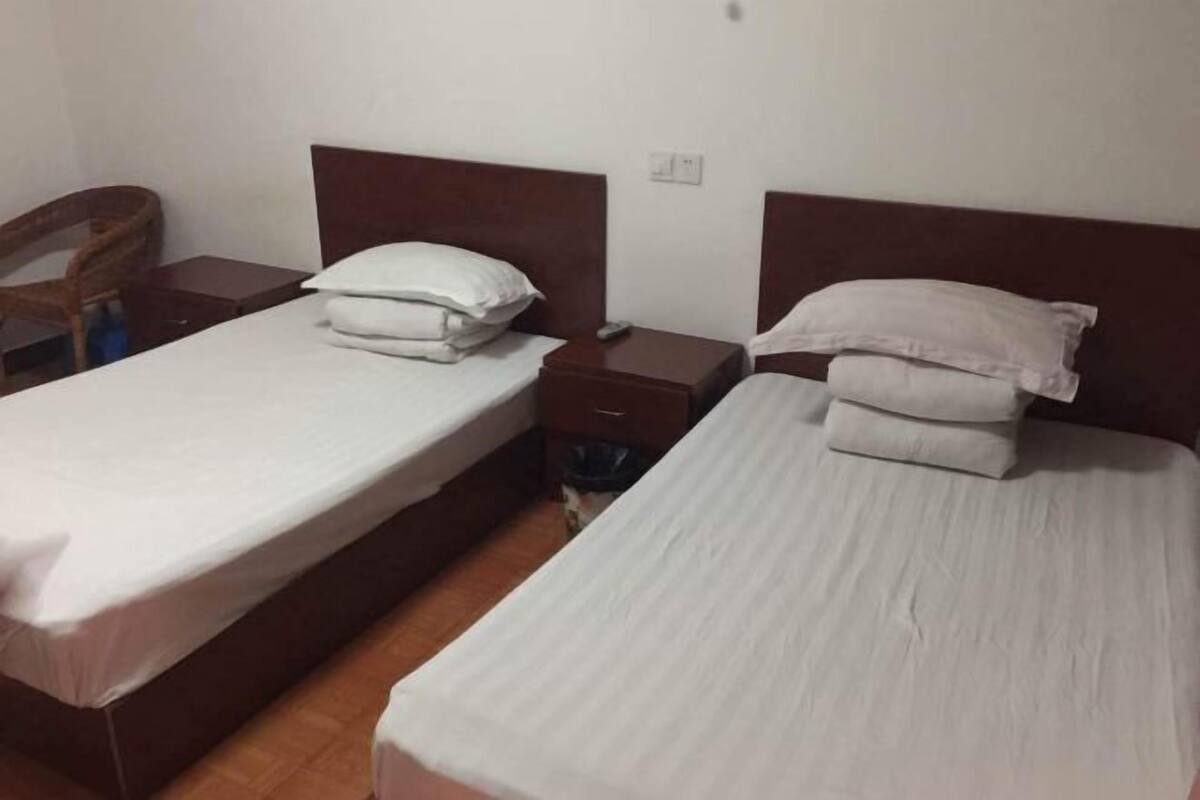 A room with two single beds, wooden headboards, a small table with a chair, and a trash bin.