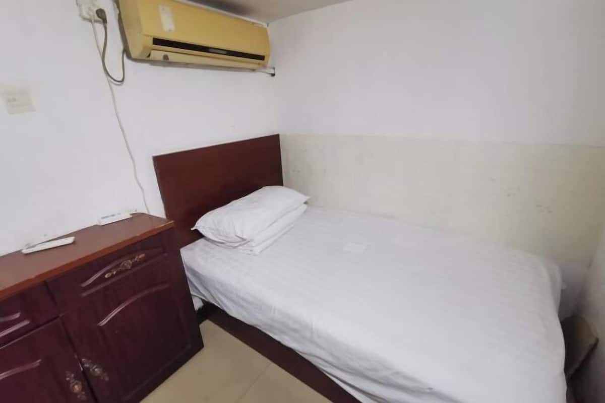 A small, simple room with a single bed, a wooden dresser, and a wall-mounted air conditioner.