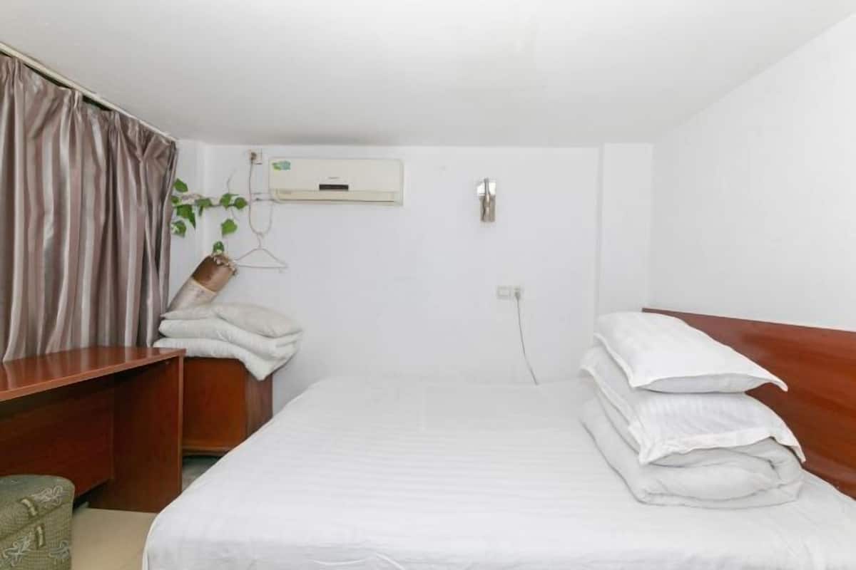 A hotel room with a bed, a wooden desk, a chair, a plant, and a wall-mounted air conditioning unit.