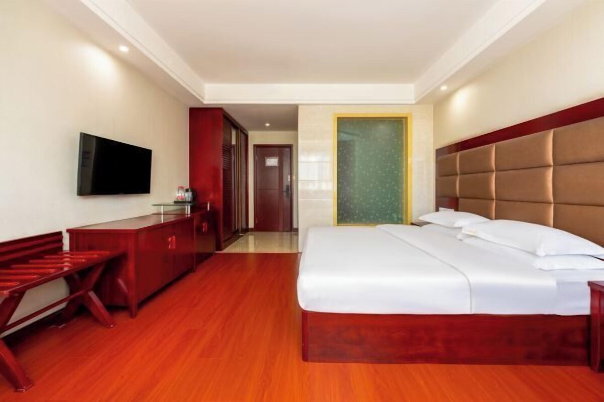 A hotel room with a large bed, a TV mounted on the wall, a desk with a chair, and a wardrobe.