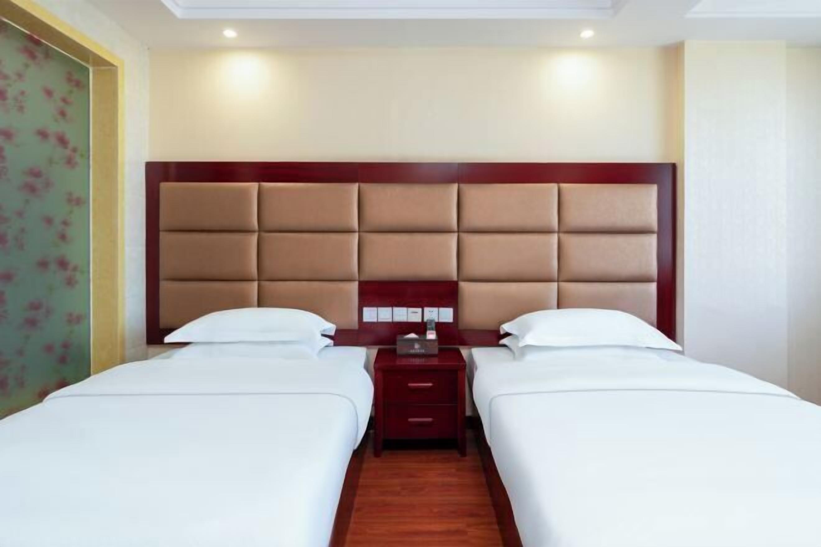 A hotel room with two beds, a wooden headboard, a nightstand, and a floral-patterned wall.