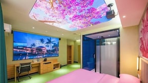 A hotel room with a large flat-screen TV, a bed with pink bedding, a desk with a chair, and a ceiling with a digital display of cherry blossoms. - Ruyi Hotel (Foshan Nanhai Songgang Avenue) (Foshan)