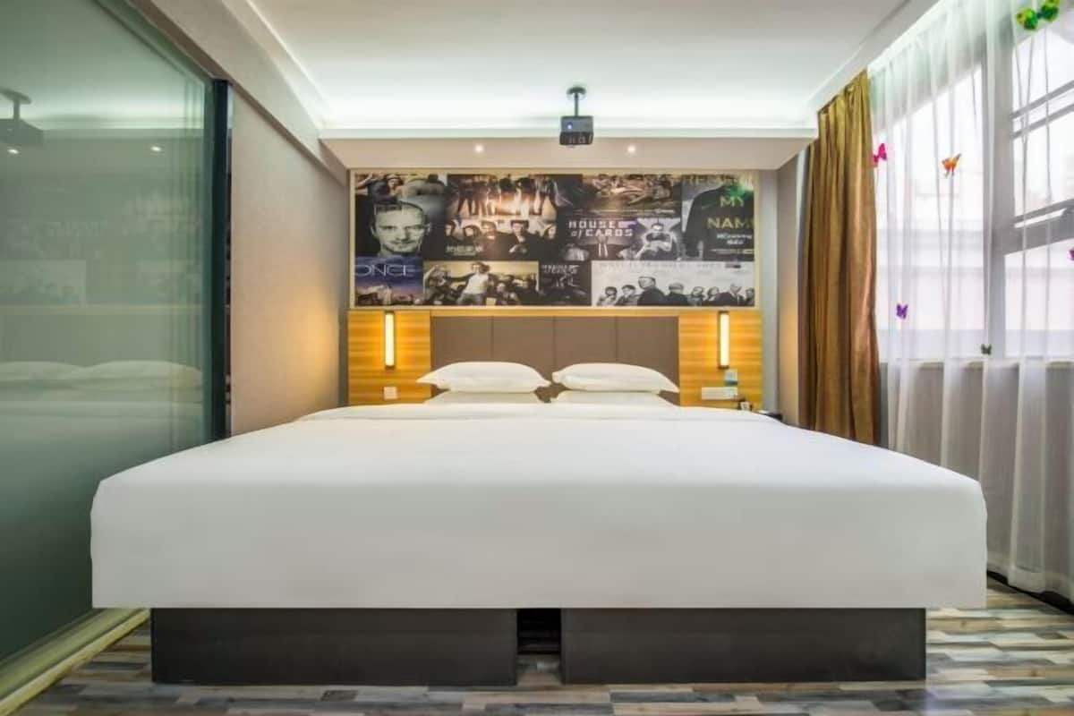 A modern hotel room with a large bed, a wall decorated with various posters, and a large window with curtains.