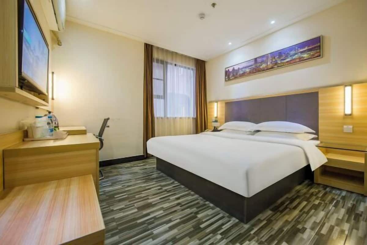 A hotel room with a large bed, bedside tables, a desk, a chair, a TV, a window with curtains, and a painting on the wall.