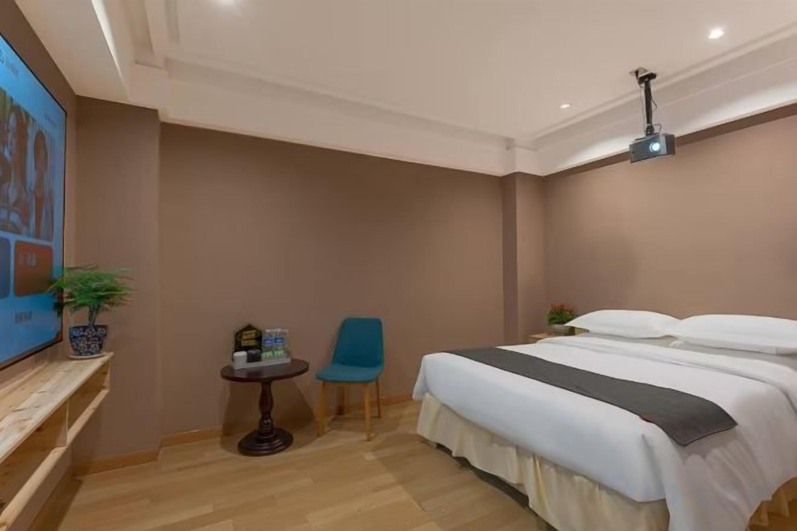 A hotel room with a bed, a television, a small table with a chair, and a ceiling-mounted projector.