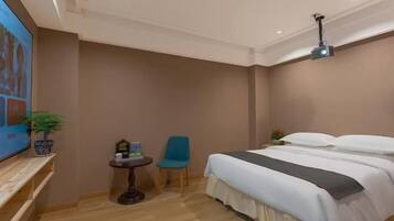 A hotel room with a bed, a television, a small table with a chair, and a ceiling-mounted projector.