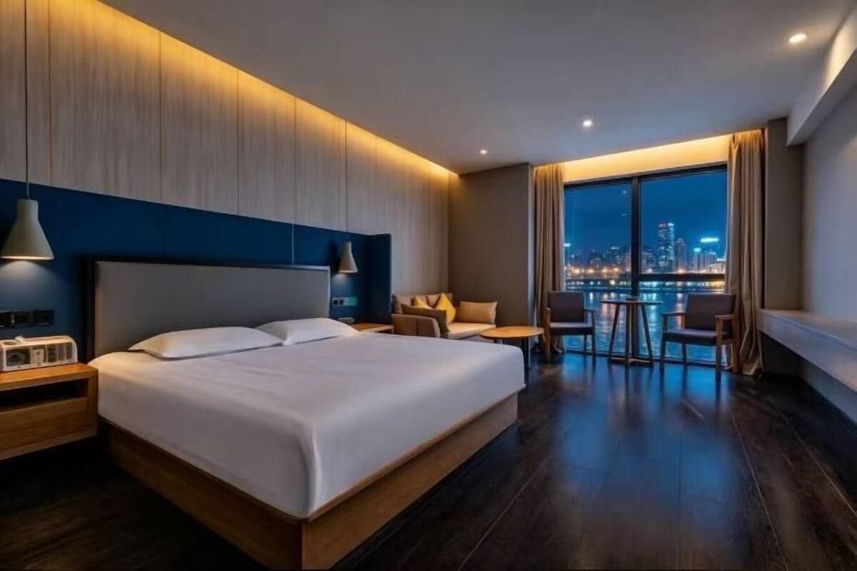 Een moderne hotelkamer met een groot bed, nachtkastjes, een zithoek met stoelen en een stadsgezicht door grote ramen.