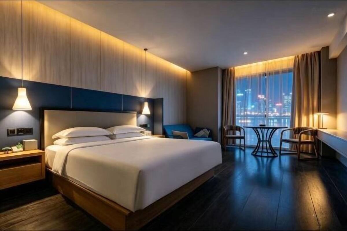 Een hotelkamer met een groot bed, nachtkastjes, een bureau met stoelen en uitzicht op de stad door het raam.