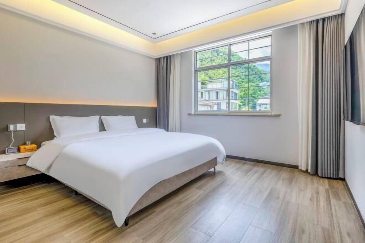 Ein modernes Hotelzimmer mit einem großen Bett, einem Nachttisch mit Lampe, einem Fenster mit Vorhängen und Blick ins Grüne.