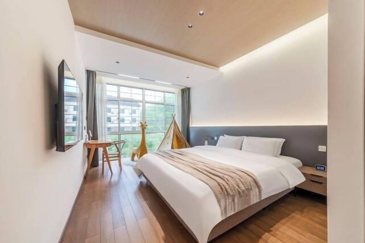 Ein modernes Hotelzimmer mit einem großen Bett, einem Schreibtisch, einem Sessel, einem Fernseher und einem Fenster mit Blick auf Gebäude.
