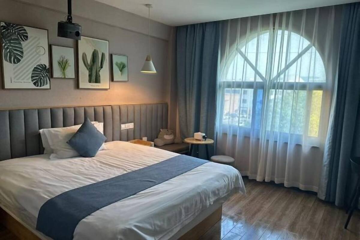 A hotel room with a bed, a small table, a chair, a window with curtains, and wall art.