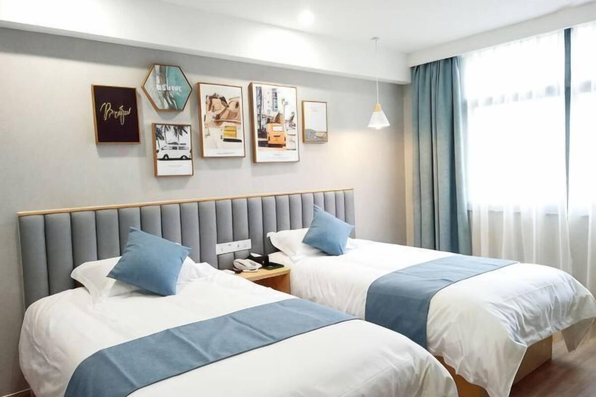 A hotel room with two beds, a grey headboard, a nightstand, and a window with curtains.