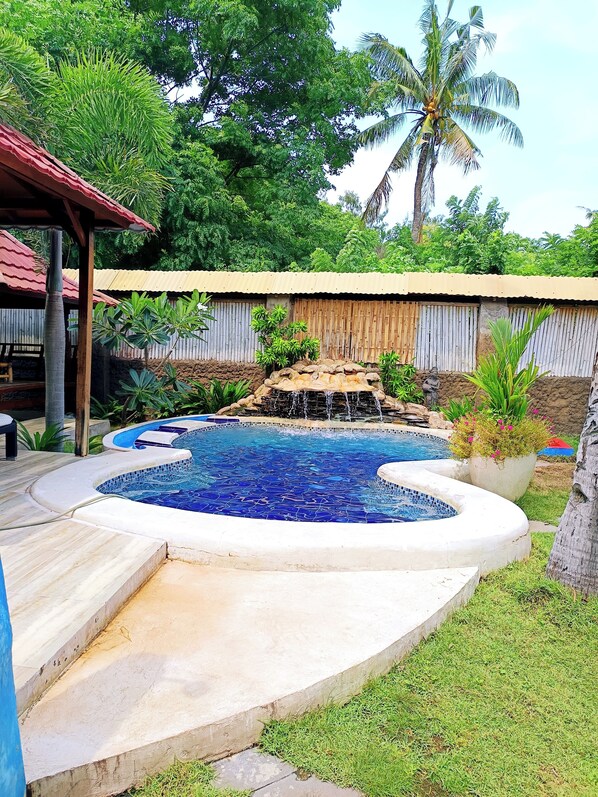 Una piscina con cascada, rodeada por una cerca de madera y vegetación tropical.