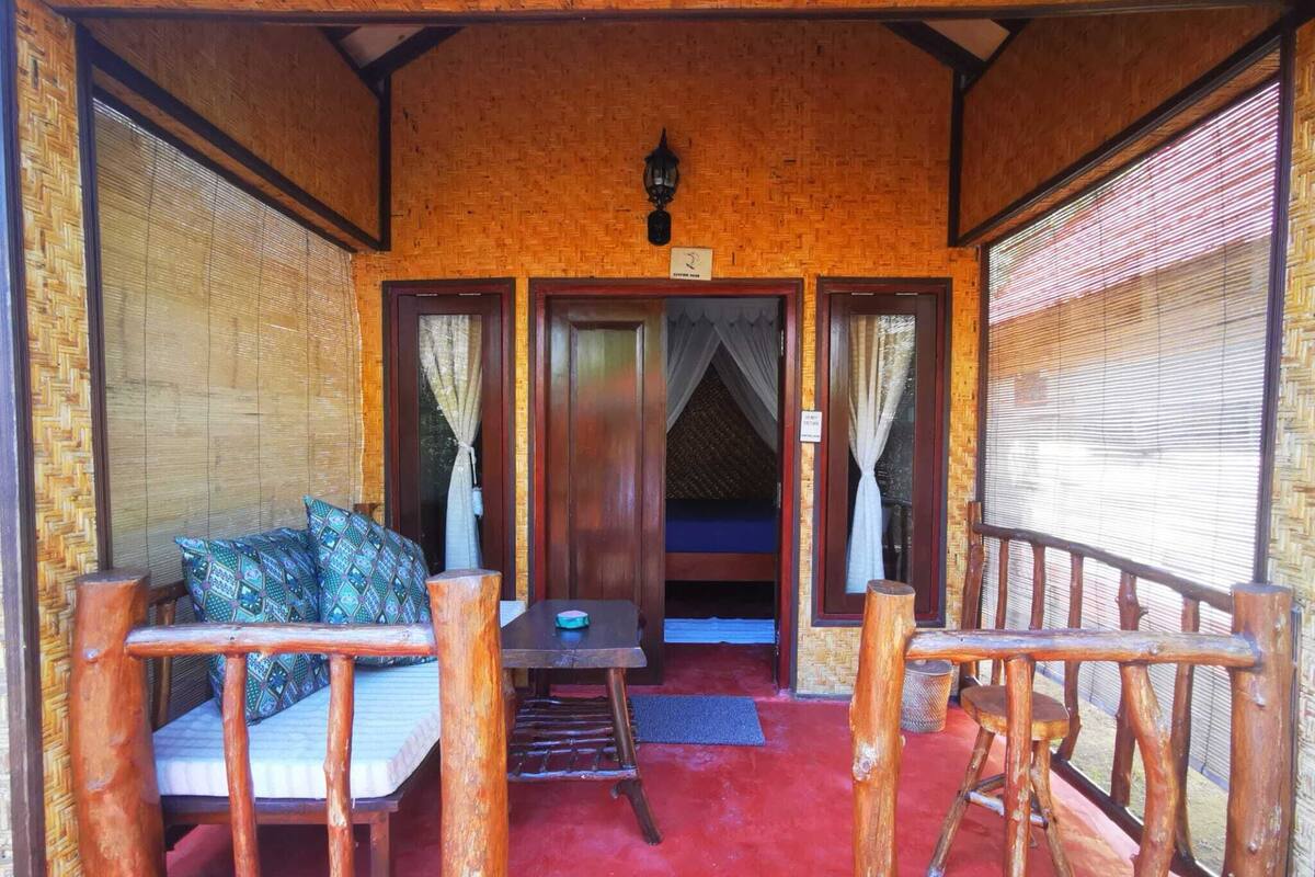 Una habitación acogedora con un banco de madera, una mesita pequeña y una cama. Hay una puerta que da a otra habitación y una ventana con cortinas.