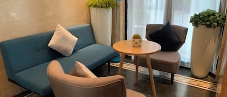 A modern lounge area with a sofa, armchair, and a small round table. There are potted plants and a lamp.