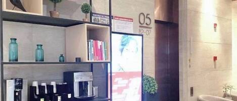 Zona de recepción de hotel con un mostrador, una pantalla digital que muestra una foto y una estantería con diversos objetos.