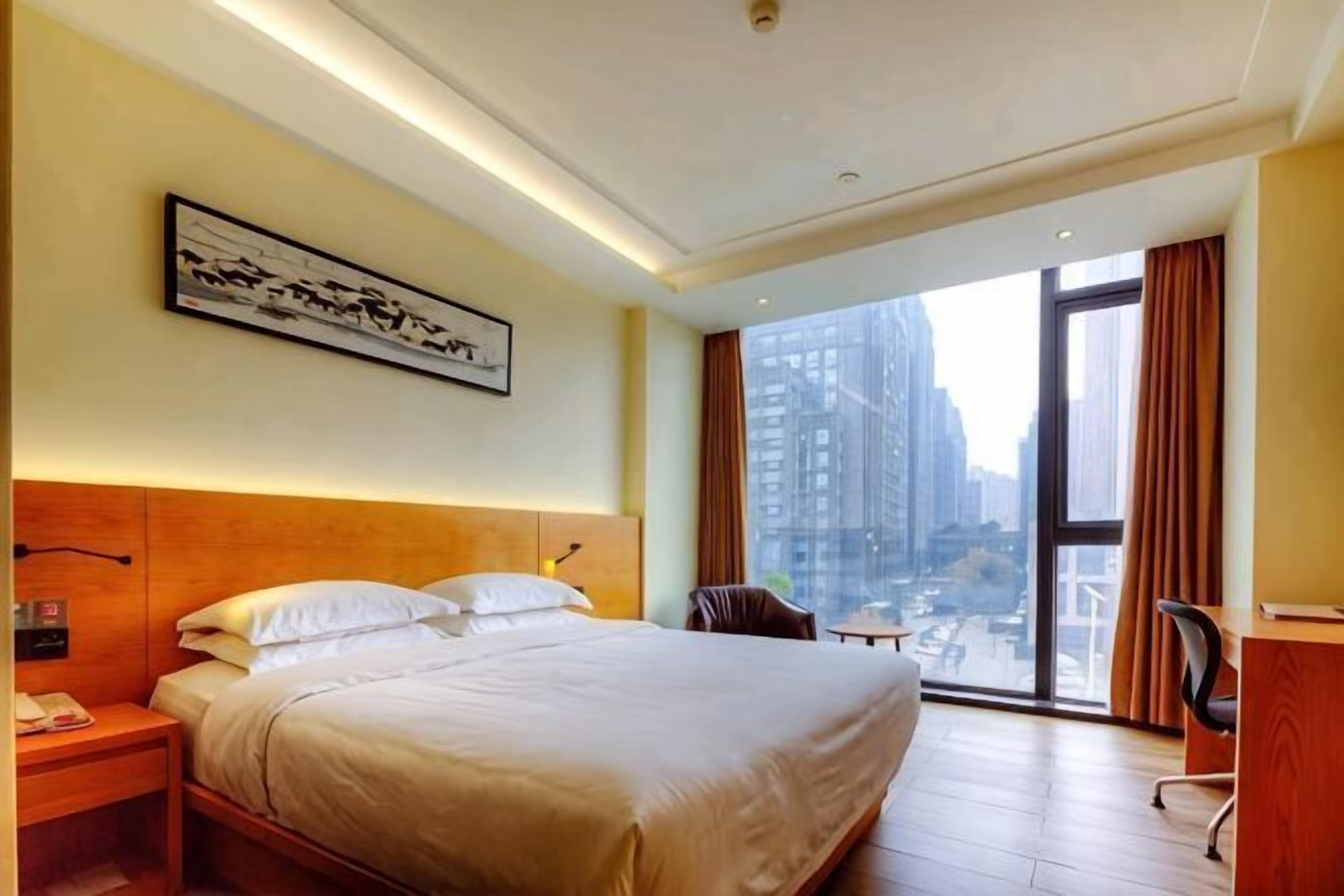 A hotel room with a large bed, a desk, a chair, a window with a city view, and a framed picture on the wall.