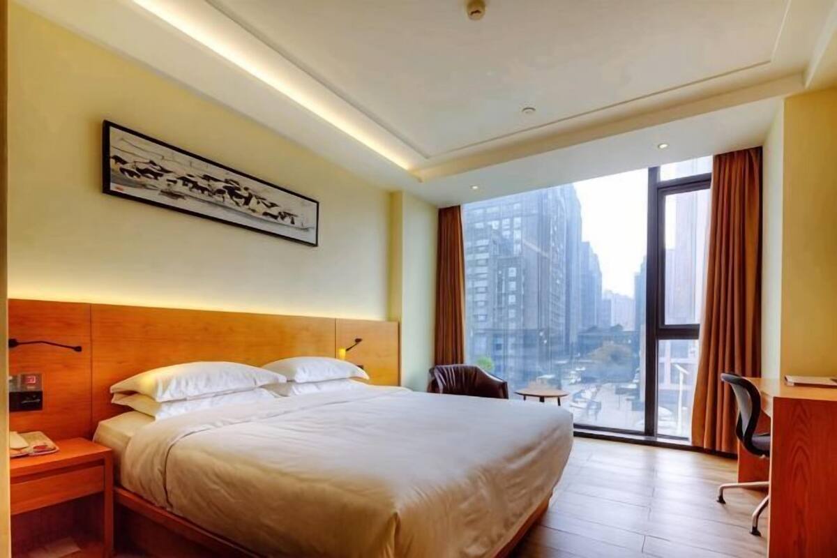 A hotel room with a large bed, a desk, a chair, a window with a city view, and a framed picture on the wall.