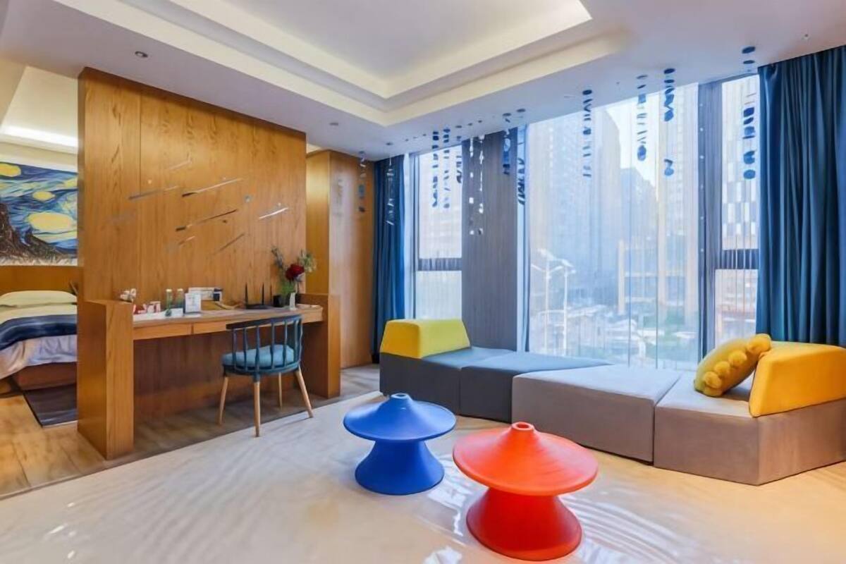 A modern hotel room with a wooden desk, a sofa, a chair, and a large window with a city view.