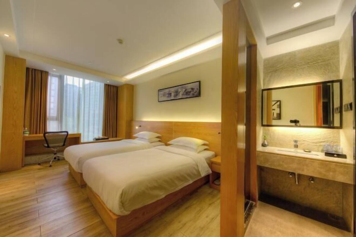 A hotel room with two beds, a desk, a chair, a large mirror, and a bathroom with a sink and mirror.