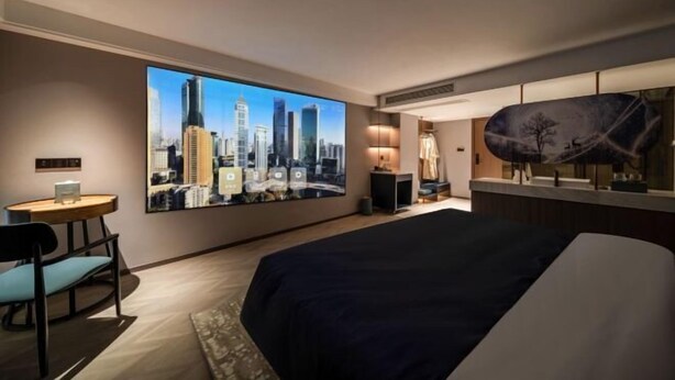A hotel room with a large cityscape painting on the wall, a bed, a desk with a chair, and a small table with a lamp. - Yearnsun Hotel (Taicang)