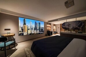 A hotel room with a large cityscape painting on the wall, a bed, a desk with a chair, and a small table with a lamp. - Yearnsun Hotel (Taicang)