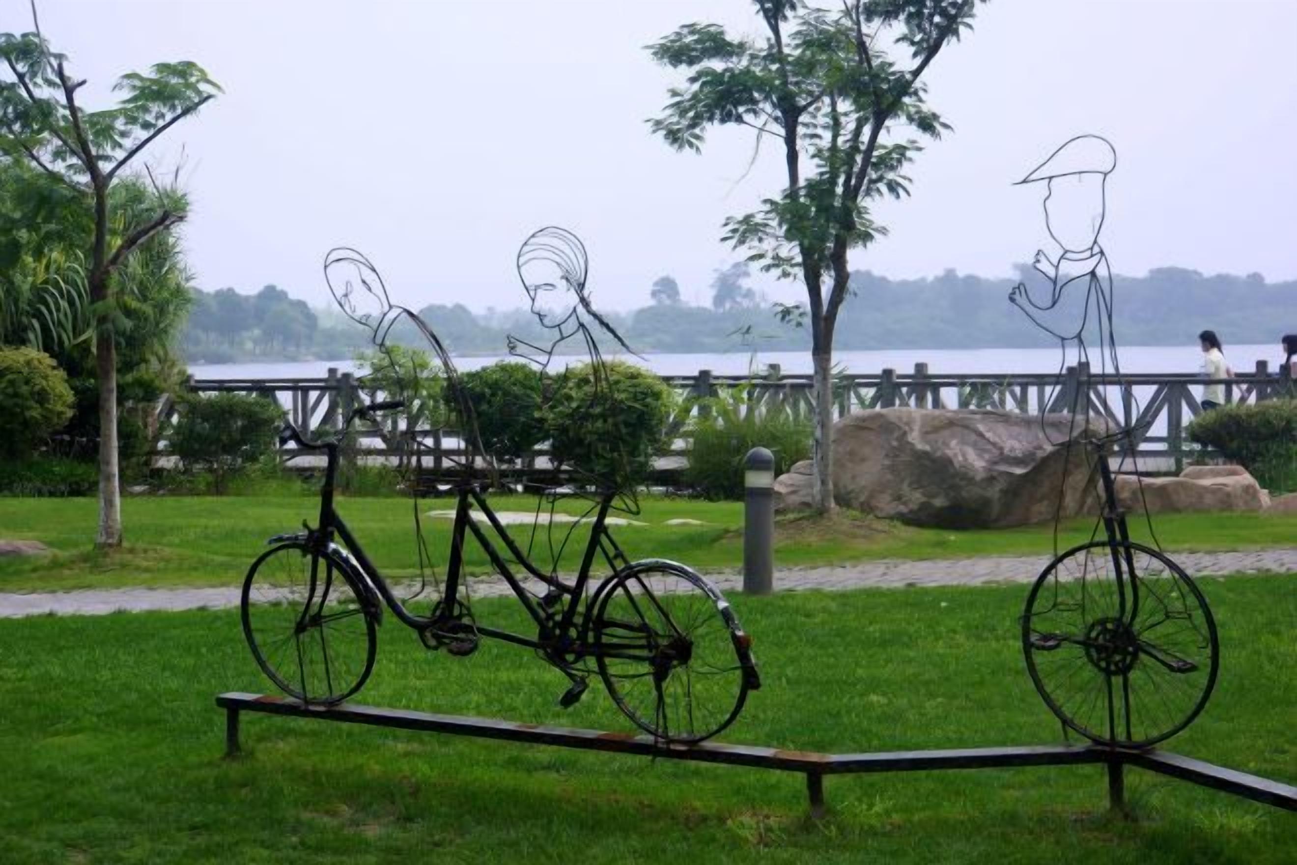 Un parque con una escultura de metal que muestra una bicicleta y una figura, un espacio cubierto de césped y un cuerpo de agua al fondo.