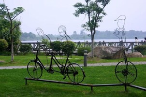 Un parque con una escultura de metal que muestra una bicicleta y una figura, un espacio cubierto de césped y un cuerpo de agua al fondo.