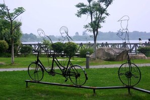 En park med en skulptur i metall som föreställer en cykel och en figur, ett grönområde och en vattenyta i bakgrunden.
