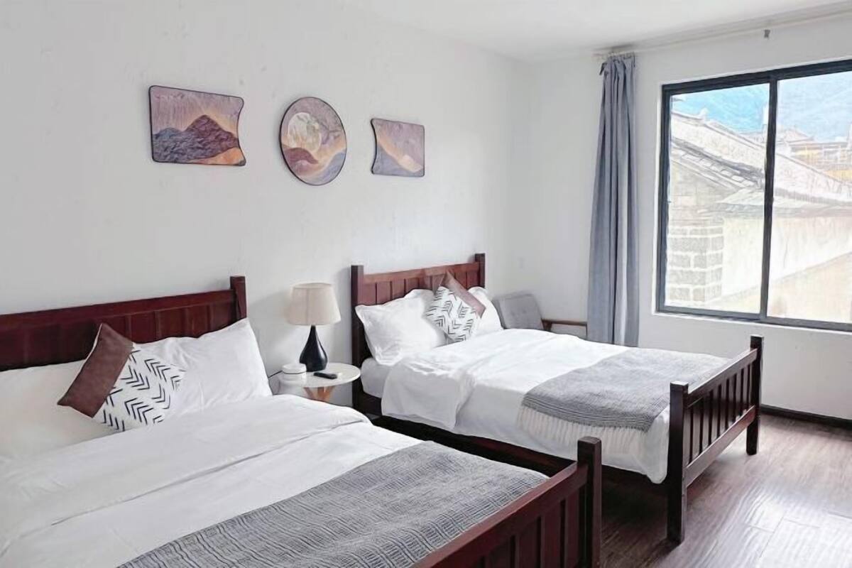 A hotel room with two beds, a nightstand with a lamp, and a window with a view of buildings.