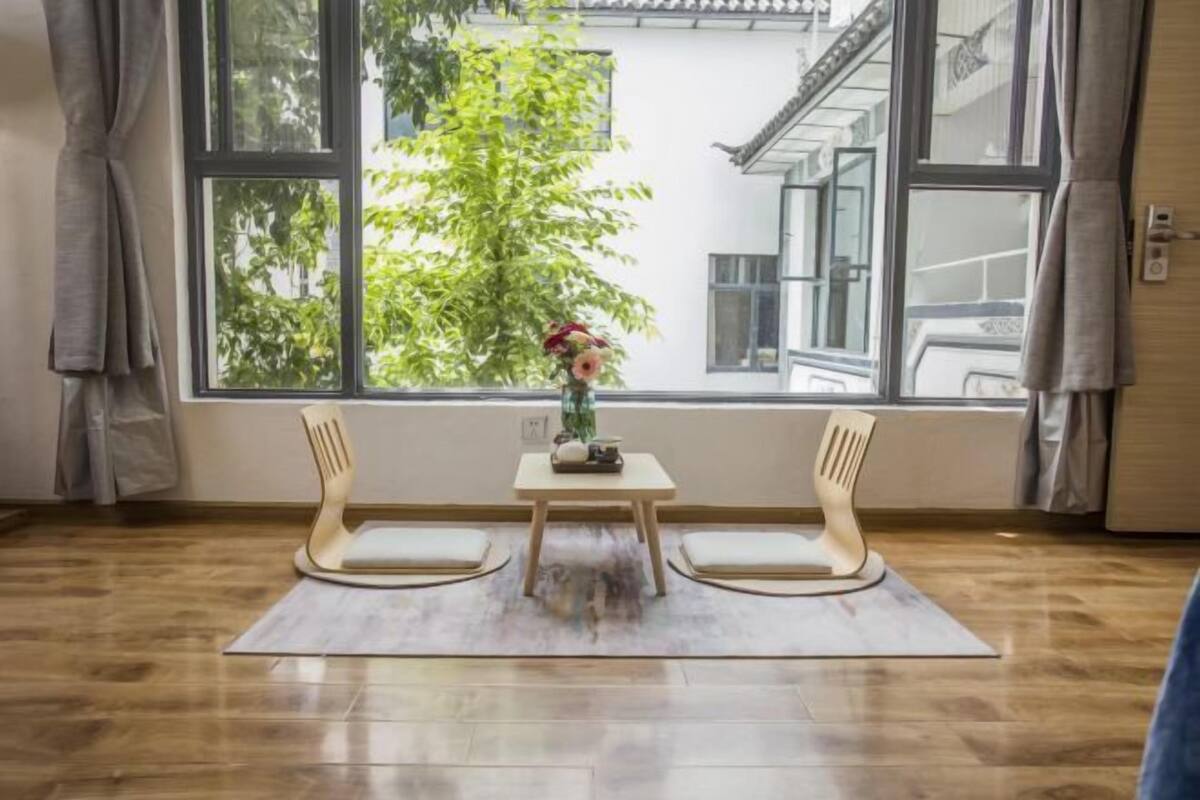 A room with a large window, a wooden table with two chairs, and a vase with flowers on the table.