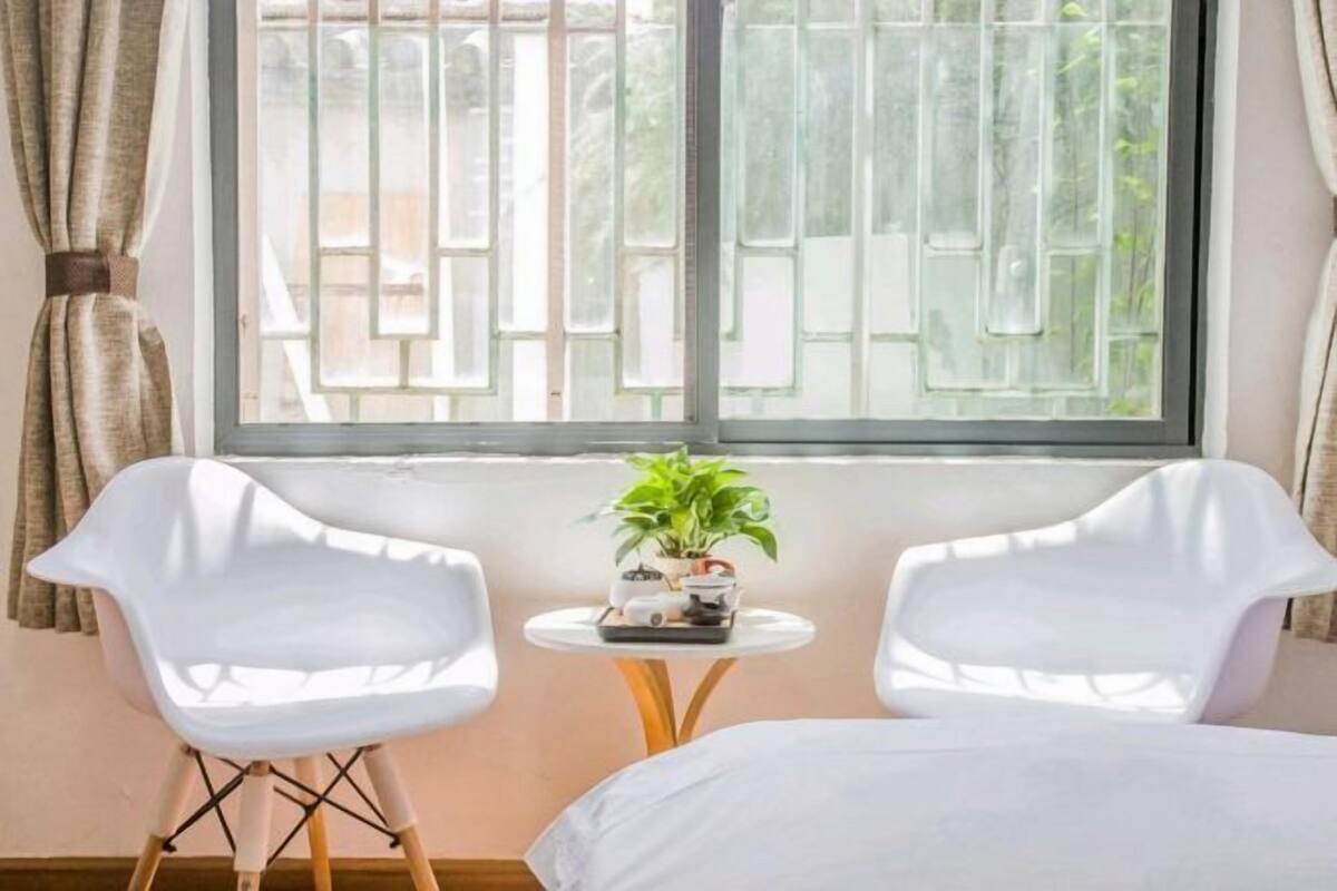 A room with a bed, two white chairs, a small table with a plant, and a window with metal bars.