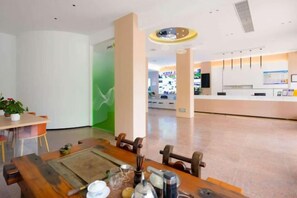 Un vestíbulo de hotel con mostrador de recepción, un área de comedor con mesa y sillas, y una pared verde con un mural de aves.