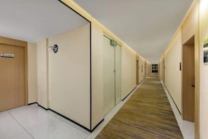 Een hotelgang met een betegelde vloer, een houten deur links, een glazen deur met een bordje en een vloerkleed.