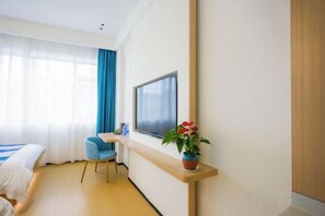 Una habitación de hotel con una cama, un televisor montado en la pared, una mesita con una planta y una ventana con cortinas.