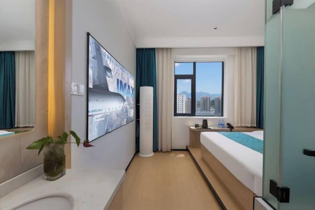 A hotel room with a bed, a window with a city view, a bedside table, a plant, and a poster on the wall.