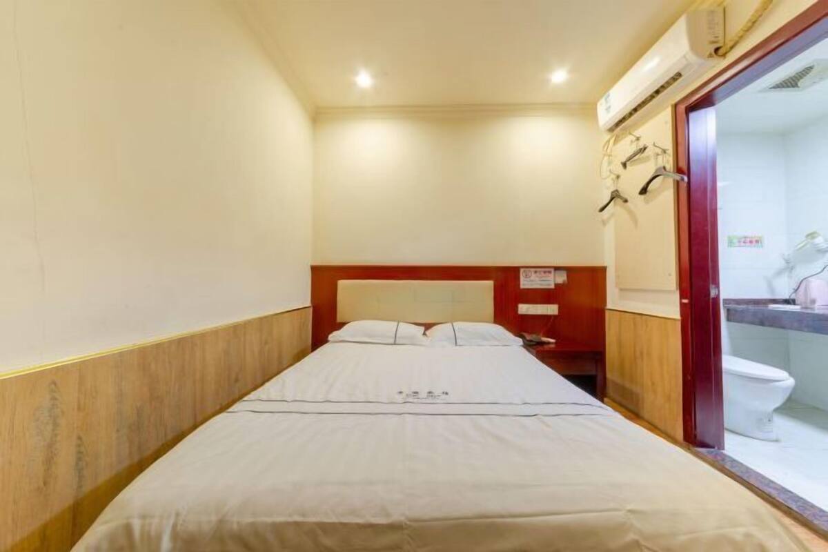 A narrow hotel room with a single bed, a wooden headboard, a small bedside table, a mirror, and a toilet visible through an open door.