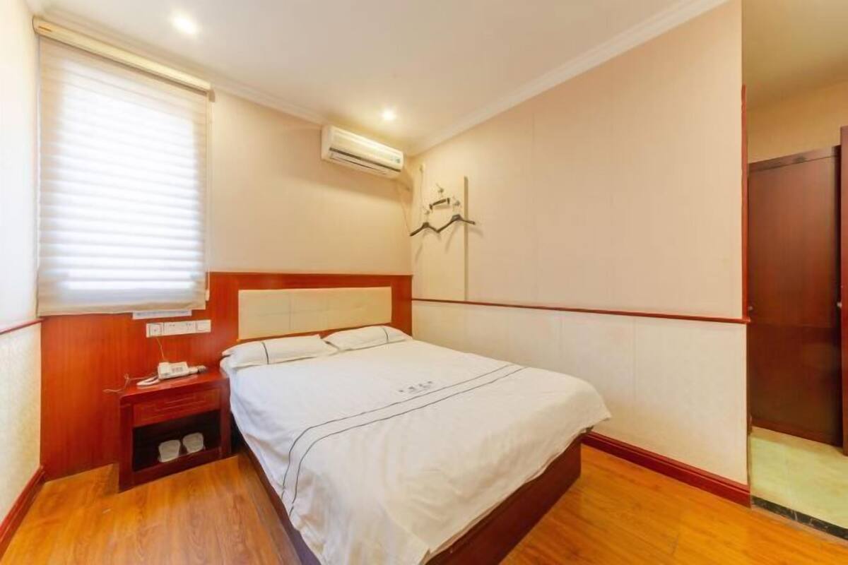 A bedroom with a wooden headboard, a bedside table with a phone, a wall-mounted air conditioner, and a door to the right.