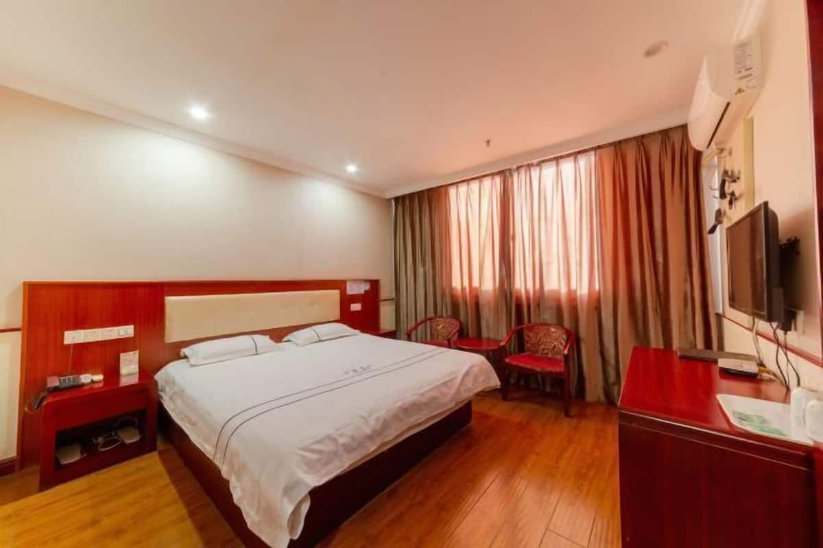 A hotel room with a wooden floor, a bed with white linens, a desk with a chair, a TV, and a window with curtains.