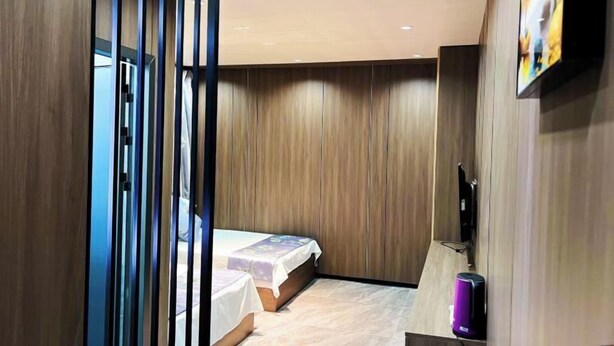 A modern hotel room with a wooden panel wall, a bed with a white bedspread, a small bedside table, and a flat-screen TV mounted on the wall.