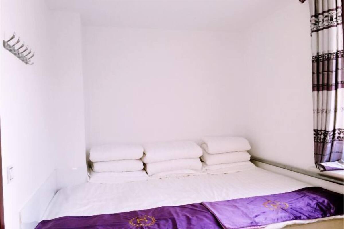 Ein Einzelbett mit weißer Bettwäsche und einer violetten Tagesdecke, ein Fenster mit gemusterten Vorhängen und ein Wandhaken.