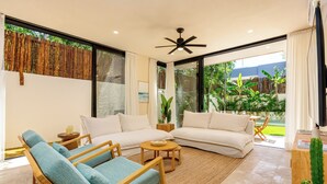 Un salon moderne avec un grand canapé, deux fauteuils, une table basse et un ventilateur de plafond. On y trouve une grande fenêtre avec des rideaux, un cactus posé sur une table en bois et une vue sur la verdure à l’extérieur.